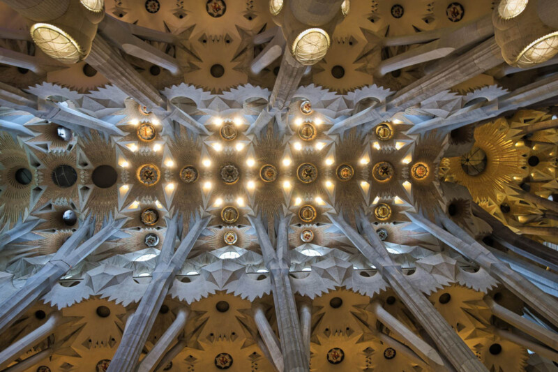 The image shows the ornate ceiling of a building, featuring a symmetrical design with star-shaped patterns and illuminated circles. Numerous columns extend toward the ceiling, adding to the intricate architectural details.