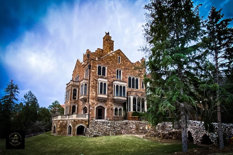 A large, historic stone mansion with multiple stories and intricate windows sits amidst a grassy area bordered by tall evergreen trees. The sky above is a vibrant, cloudy blue, enhancing the dramatic setting.