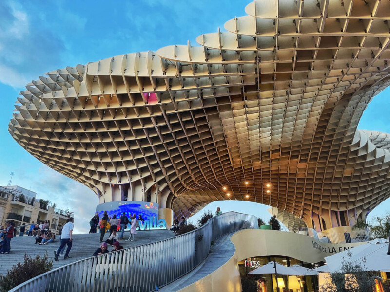 People gather around the Metropol Parasol in Seville, Spain, a wooden structure with a lattice design. The view showcases its undulating form against a blue sky, with stairs and pathways bustling with visitors below.