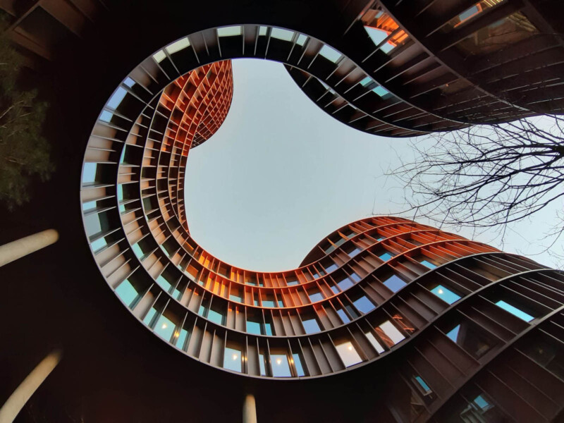 A view looking up at a modern, curved building with reflective glass windows and warm exterior tones. The shape creates an organic, flowing form against a clear sky, with a tree branch visible on the right.