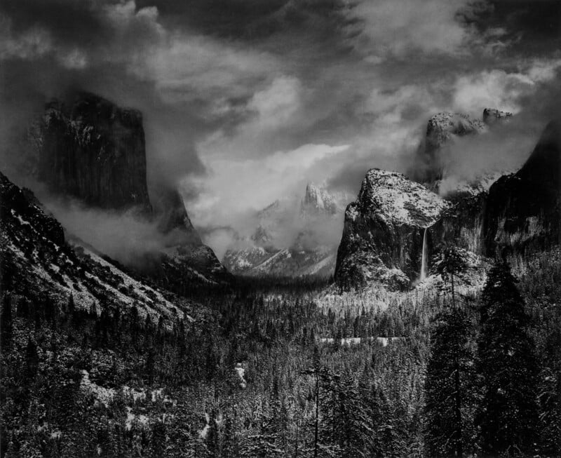 A black and white photograph of a misty Yosemite Valley, featuring dramatic cliffs and snowy forests. Clouds hang low over the landscape, partially obscuring the mountains, conveying a serene and mysterious atmosphere.