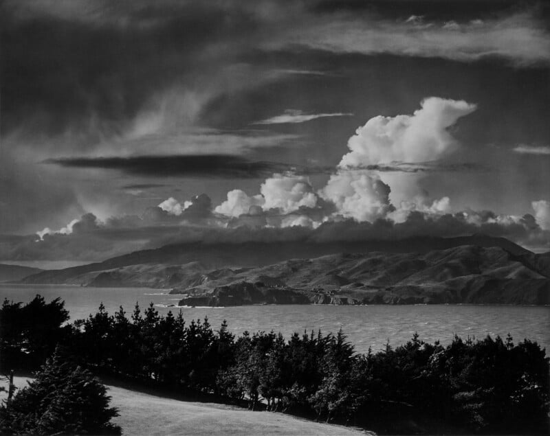 A black and white landscape photograph showing dramatic clouds over a mountainous coastline. The foreground features a line of trees adjacent to a calm body of water, reflecting light from the cloud-filled sky.