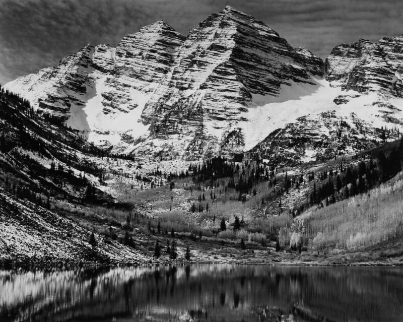 A black and white image of snow-capped mountains reflected in a calm lake. The rugged peaks tower above a forested landscape, with patches of snow visible on the slopes and water. The sky is cloudy, enhancing the dramatic scenery.