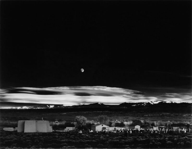 Black and white image of a landscape with distant mountains under a dark sky. A small, dimly lit town is visible in the foreground, with a bright moon shining above, partially obscured by clouds.