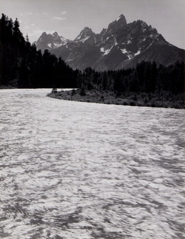 Photographie en noir et blanc d'une large rivière qui coule à travers une zone boisée, avec des sommets montagneux escarpés et enneigés en arrière-plan. Les arbres et les montagnes créent un contraste saisissant avec l’eau brillante et chatoyante.