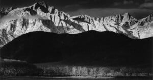 Black and white landscape of jagged, snow-capped mountains contrasting against dark rolling hills. Leafless trees line the foreground, while a lone figure on horseback is visible below the hills.