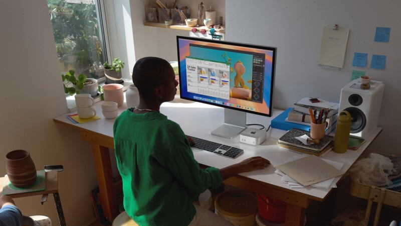 A person sits at a desk working on a computer in a bright room. The desk has pottery materials, a speaker, and a notebook. There are plants on the windowsill and art supplies on shelves. The screen displays a creative project.