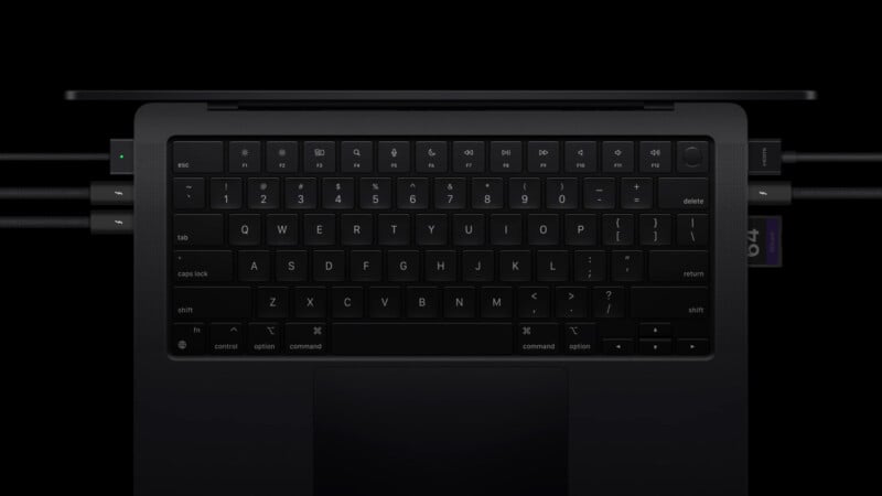 A close-up shot of a laptop keyboard, featuring a black QWERTY layout. The screen is not visible, and the keys are illuminated subtly, with cables connected on the left side. The background is dark, emphasizing the keyboard's sleek design.