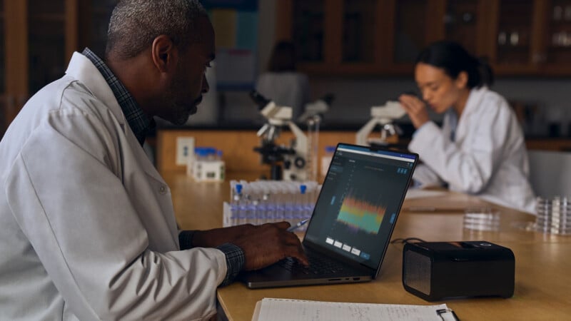 A scientist in a lab coat is working on a laptop at a laboratory table, with test tubes and a small electronic device nearby. Another scientist in the background examines a sample under a microscope. Shelves filled with equipment are visible.