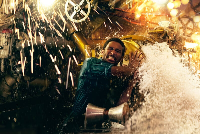 A person in a blue shirt struggles to turn a valve amid a burst of water and flying sparks in a dimly lit industrial setting. The intense scene conveys urgency and effort in managing the situation.