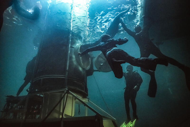 Divers swim underwater near a large cylindrical structure, illuminated by blue light. Some wear fins and masks, creating bubbles as they move. The atmosphere is mysterious and deep, with shadows and reflections on the water surface.