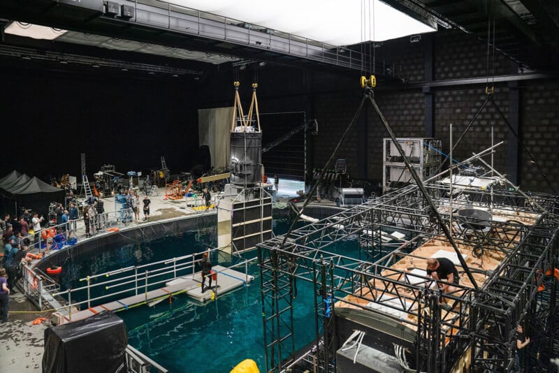 Film set featuring a large indoor water tank surrounded by various equipment and crew members. There are scaffolds, cameras, and lighting rigs around the pool. People are working on and around the water, indicating an active scene in production.