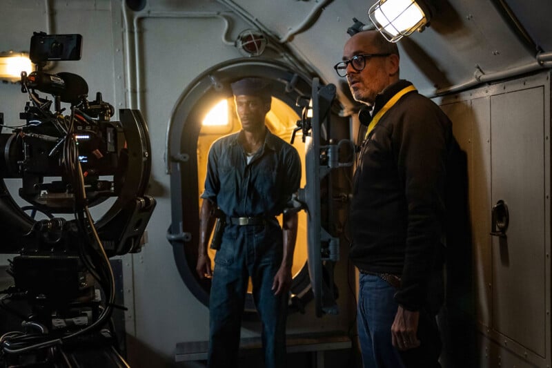 A man wearing a cap stands in the doorway of a dimly lit submarine interior, while another man in glasses and a dark shirt stands nearby, next to filming equipment and cameras. They appear to be on a film set.