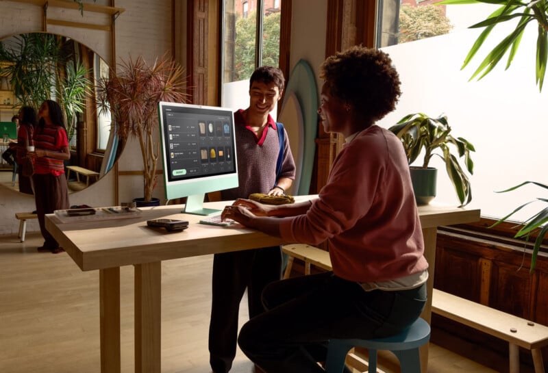 Two people are interacting at a standing desk with a large monitor displaying an app interface. The room is filled with plants, natural light, and features wooden floors. A person in the background is on their phone.