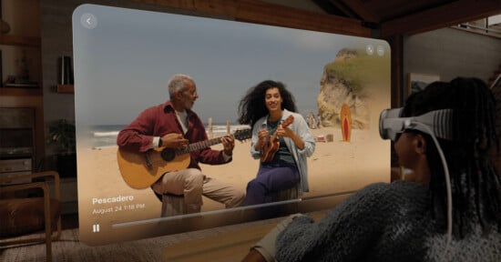 A person wearing virtual reality glasses watches a video of an older man and a young woman playing guitars on a beach. The scene is labeled "Pescadero" with a date and time. A surfboard rests against a rock in the background.