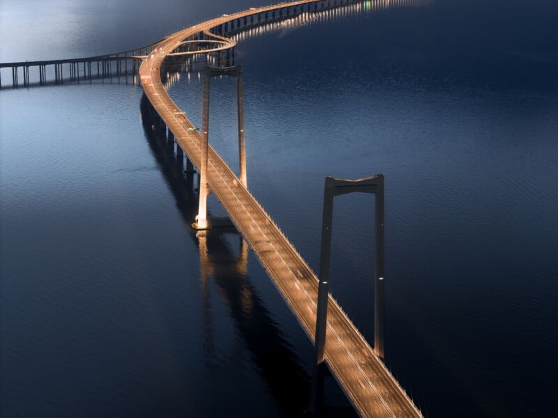 Aerial view of a long, curved bridge spanning across calm blue water, illuminated by streetlights. The bridge's golden glow contrasts with the dark, serene surface of the water below.