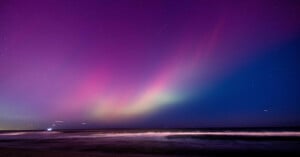 A mesmerizing sky displaying a gradient of purple, pink, and green auroras over a dark ocean at night. Gentle waves brush the shore under a starry sky, creating a serene and captivating scene.