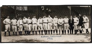 A vintage black and white photo of a baseball team from 1914. The players are lined up on a baseball field wearing uniforms, with some holding bats. "Baltimore International 1914" is written at the bottom. The background shows empty bleachers.