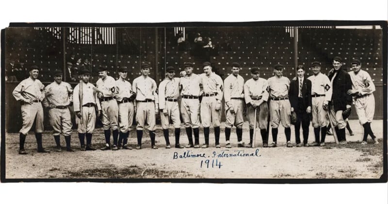 A vintage black and white photo of a baseball team from 1914. The players line up on a baseball field, dressed in uniforms, some holding bats. "Baltimore International 1914" is written at the bottom. Empty stands can be seen in the background.