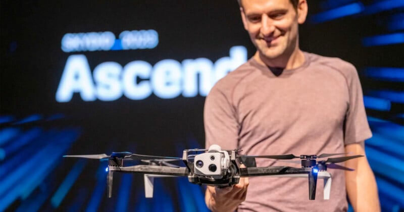 A man smiles while presenting a drone in front of a blurred background that reads "Ascend." He wears a casual shirt, and the drone is featured prominently in the foreground.