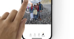 A hand uses a phone app to edit a photo of a family standing on a rocky beach with cliffs in the background. App options displayed include Adjust, Filters, Crop, and Clean Up.