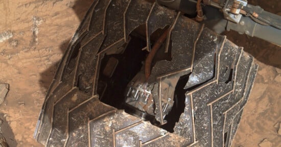 A close-up of a damaged space rover wheel on a dusty surface, showing a large crack and missing segment. The wheel's tread pattern is visible, and a portion of the rover's mechanism can be seen through the gap.