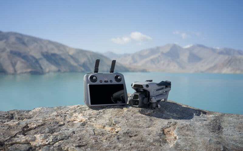 A drone and its controller are placed on a rocky surface with a scenic backdrop of a tranquil lake and distant mountains under a clear blue sky.