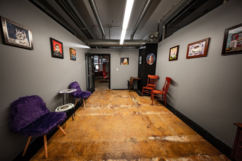 A narrow hallway with gray walls, decorated with various framed artworks. Two purple fur-covered chairs with small tables face each other on the left, and two wooden chairs are on the right. The floor is worn and there is a doorway at the end.