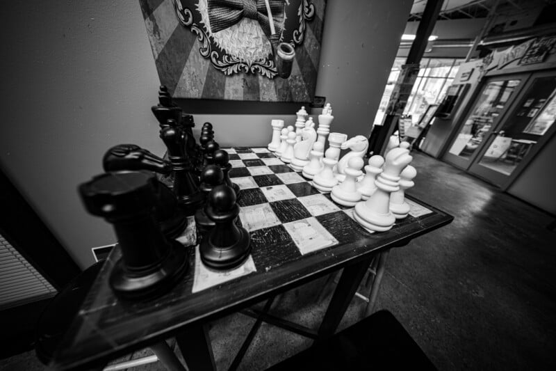 Black and white image of a large chessboard with oversized chess pieces set up for a game. The scene is indoors, with artwork and a tiled floor visible in the background.