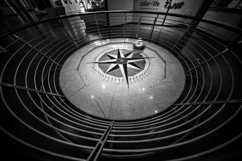 A black and white image of a spiral staircase with a railing encircling a large floor compass at the center. The compass design features directional points, and the scene is viewed from above.