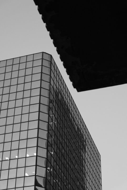 Black and white image of a modern glass building, reflecting another structure. The building features a grid of reflective windows, with part of a dark overhang or roof visible in the upper right corner against a clear sky.