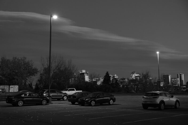 Schwarz-Weiß-Bild eines Parkplatzes mit geparkten Autos, beleuchtet von Straßenlaternen. Im Hintergrund ist die Skyline einer Stadt unter einer dramatischen Wolkenformation am Abendhimmel zu sehen.