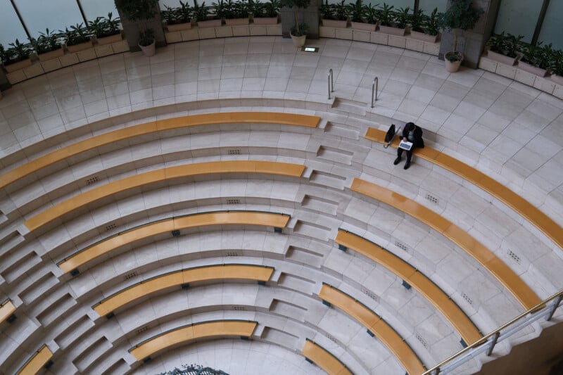 A person sits alone on yellow benches in a spacious, circular auditorium. The seating is arranged in curved rows with light-colored flooring and potted plants lining the perimeter.