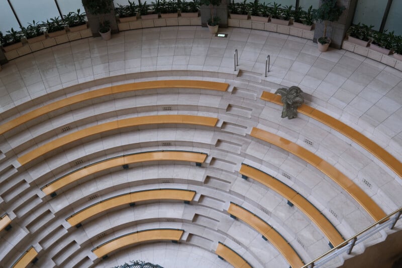 Aerial view of a circular, open-air amphitheater with yellow seating. The seating is arranged in tiers around a central performance area. Potted plants adorn the upper perimeter, and a lone statue sits among the seats.