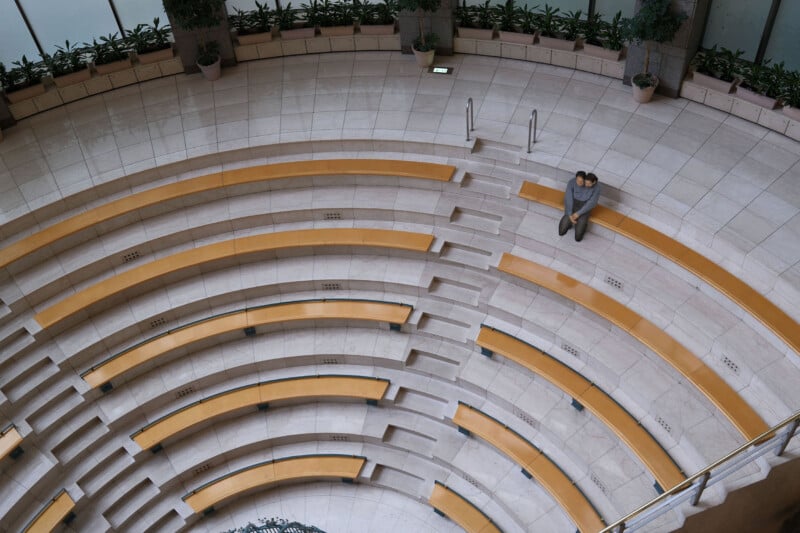 Aerial view of an empty amphitheater with curved yellow benches. A person in gray sits alone on one of the benches, surrounded by potted plants along the perimeter. The setting appears indoors with natural lighting.