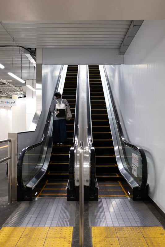 A person wearing a mask stands on an escalator at a train station, holding a bag and phone. The station features bright lighting, metallic and white walls, and safety markings on the floor.