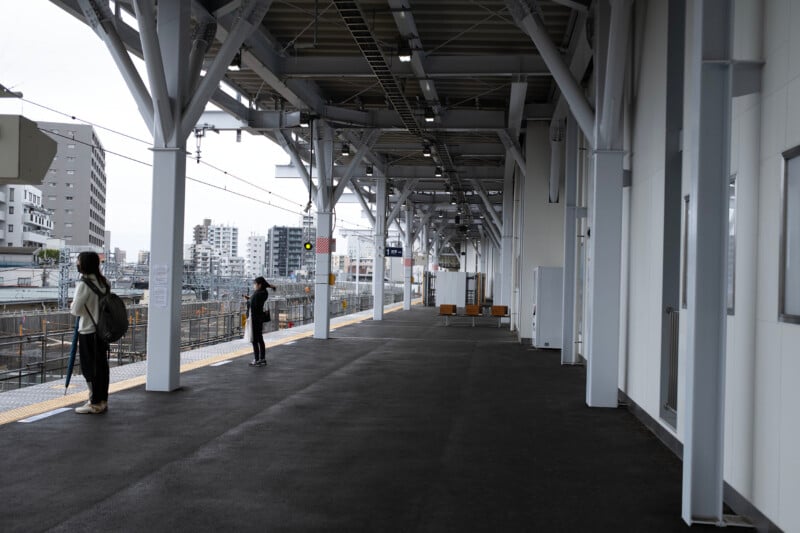 A nearly empty train platform under a modern, white steel structure. Two people stand near the edge, facing the tracks. In the background, urban buildings are visible against a cloudy sky. Several benches are unoccupied.