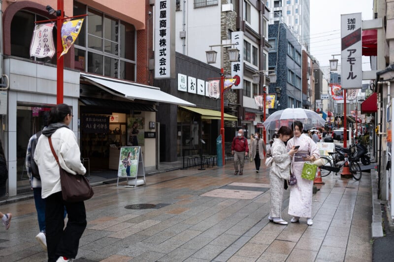 People walk along a rainy street in a shopping area. Two individuals in traditional clothing hold an umbrella. Various shops and signs line the street, with some pedestrians carrying umbrellas.