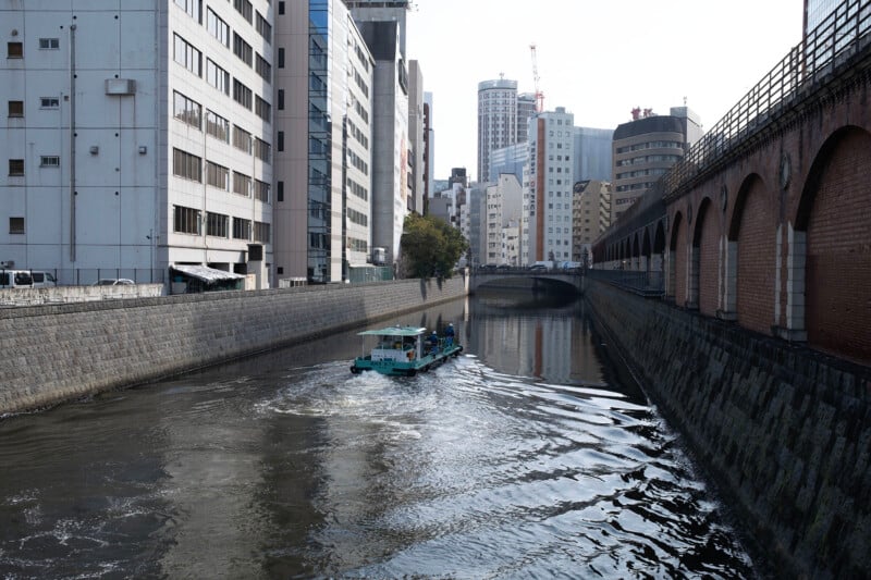 A small boat travels along a canal bordered by modern buildings and a brick wall on one side, reflecting in the water. The cityscape and overcast sky create an urban atmosphere.