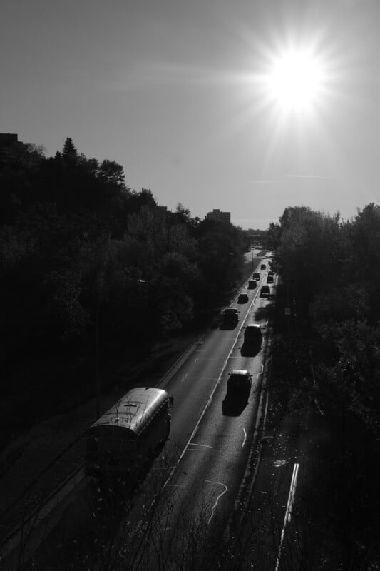 Schwarz-Weiß-Bild einer Straße mit Fahrzeugen, die sich bei strahlendem Sonnenschein in beide Richtungen bewegen. Bäume säumen die Straße und die Landschaft ist leicht hügelig. Das Sonnenlicht erzeugt starke Schatten und Glanzlichter auf der Szene.