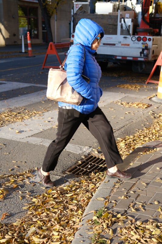 Eine Person, die eine leuchtend blaue Pufferjacke und eine Sonnenbrille trägt, geht über eine mit Blättern bedeckte Straße. Sie tragen eine beige Tasche und betreten einen Bordstein. Im Hintergrund sind ein LKW und orangefarbene Baukegel zu sehen.