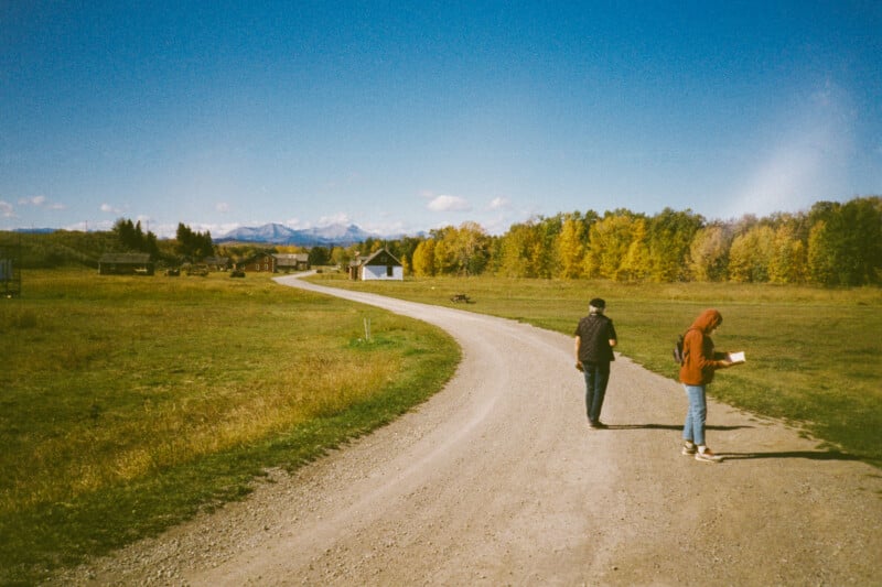 A winding dirt road leads through a grassy field toward a cluster of houses and distant mountains. Two people walk along the road under a clear blue sky. Autumn trees line the right side, with vibrant yellow leaves.