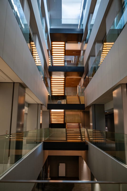 Modern multi-story building interior with illuminated staircases and glass railings. The architecture features clean lines and an open design, with natural light streaming in from overhead windows.