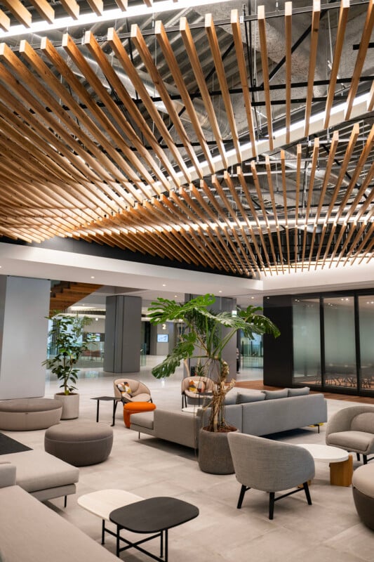 Modern lobby with gray seating, round poufs, and a central plant. The ceiling features wooden slats and linear lighting. The space has a minimalistic design with a mix of neutral tones and a few pops of orange.