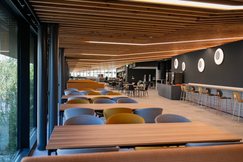 A modern cafeteria with wooden tables and a mix of yellow and gray chairs. The ceiling has wooden slats with embedded lighting. The far wall is dark with round light fixtures, and a counter area with high stools is visible in the background.