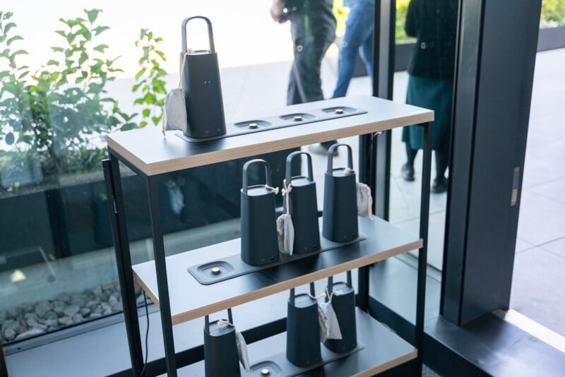 A modern black wooden shelf holds several cone-shaped portable lanterns with handles, each resting on a circular charging base. The setup is next to a glass door with outdoor foliage visible.