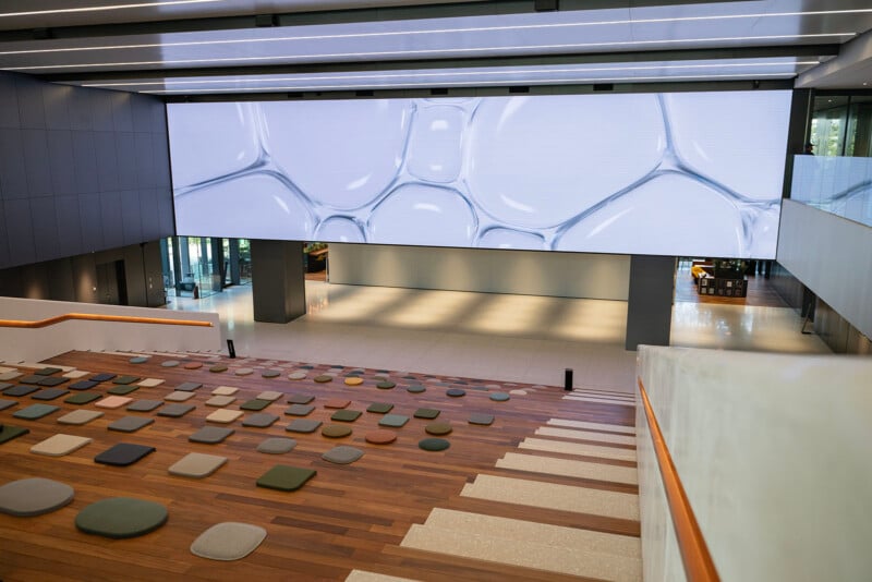 A modern interior with tiered wooden seating and small colorful cushions. A large digital screen displaying abstract patterns dominates the wall. The space is open and bright, with sleek design elements.