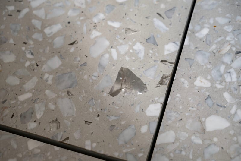 Close-up of a terrazzo floor with a chip in one tile. The surface features a mix of light gray, dark gray, and white stone chips embedded in a gray cement base, highlighting the damage.