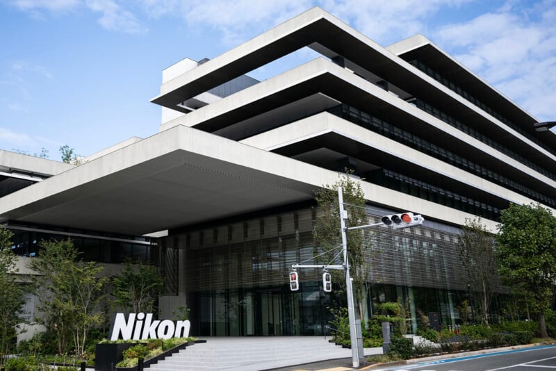 Modern building with a sleek, tiered design and large glass windows, surrounded by greenery. The Nikon logo is prominently displayed near the entrance. Cloudy blue sky in the background.