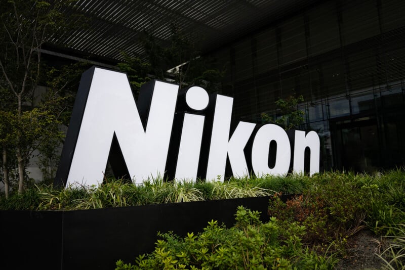 Large white 3D "Nikon" logo displayed outdoors, surrounded by greenery. The sign is set against a modern building with glass windows and a metal roof structure overhead.